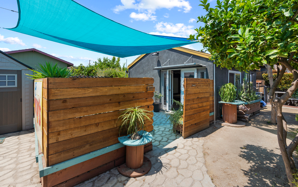Backyard detached ADU with a private patio entrance, wood privacy fence, shade sail canopy, and landscaped garden area.