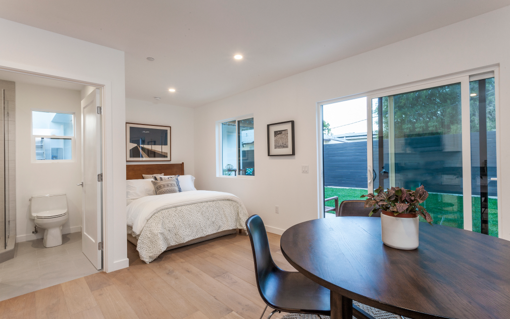 Bright ADU interior with open studio layout, dining table, bed area, sliding glass doors to backyard, and bathroom with shower.